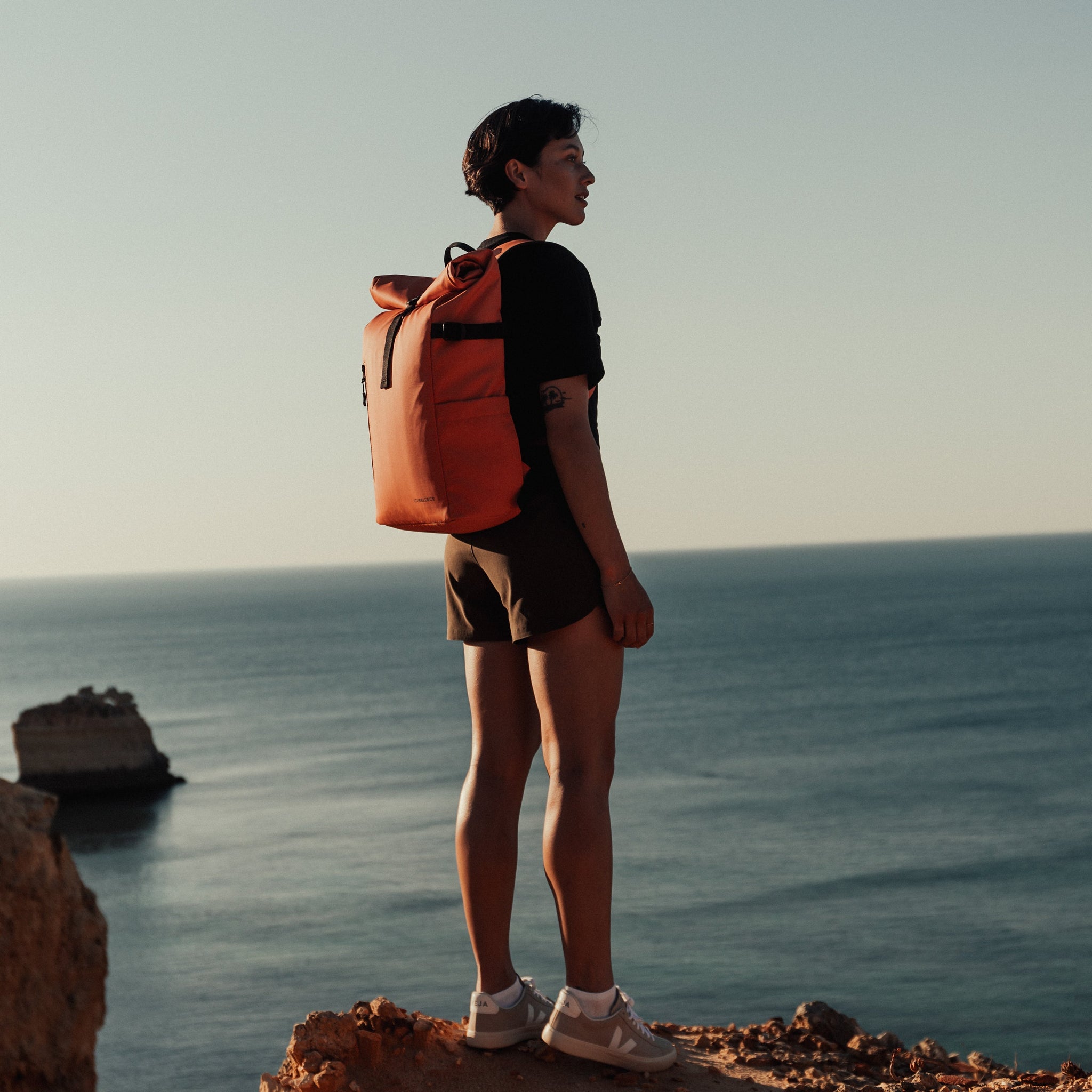 A women standing on a rock on a beach wearing The Roll Top in Ember Orange