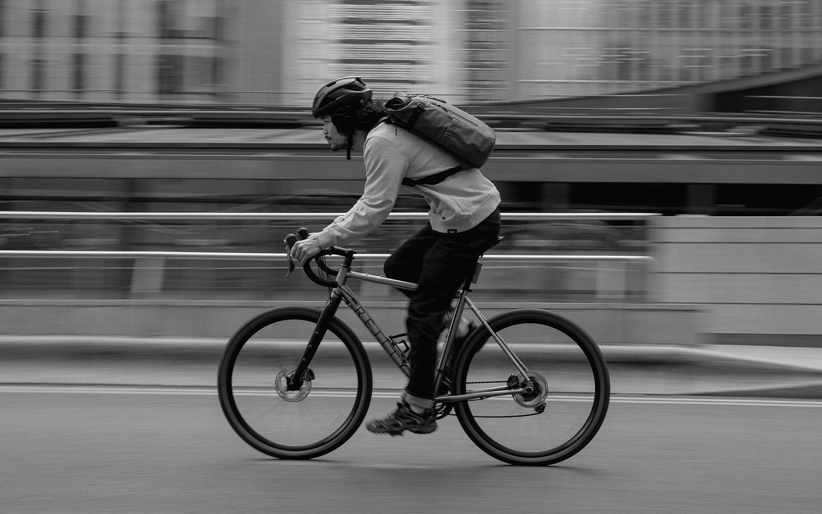 A black and white shot of a man cycling with a roll top 20l on his back