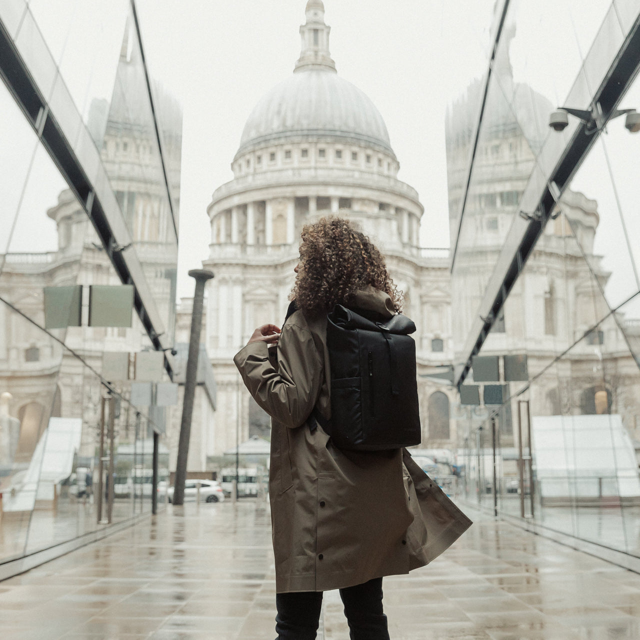 Women in a rain coat stood on a London street, with her back to the camera wearing The Roll Top Mini in All Black.