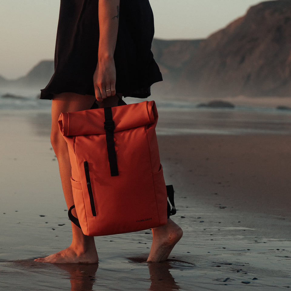 A woman walking on the beach holding a Roll Top Mini backpack in Ember Orange by her side