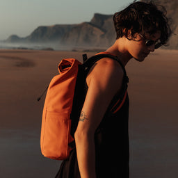  A women with Roll Top Mini backpack in Ember Orange on her back standing on the beach in the sunset