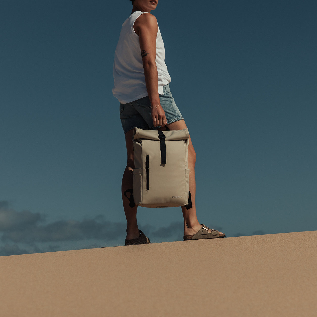 A woman standing on the beach holding Roll Top Mini backpack in Sand