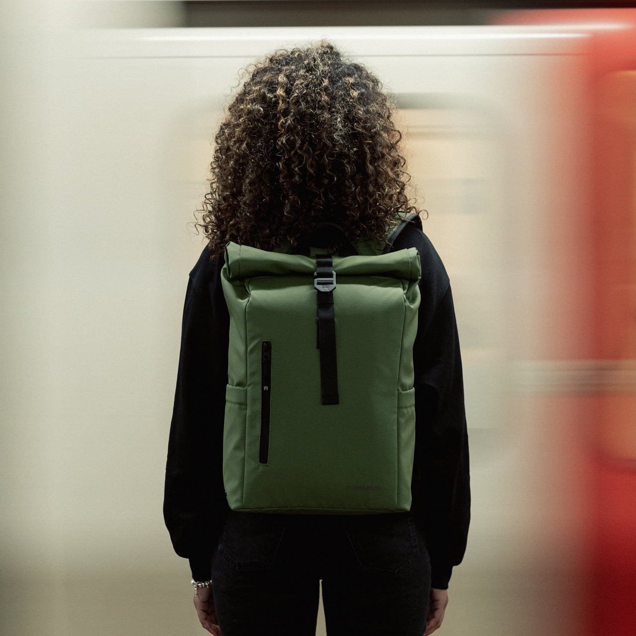 A woman standing in front of a tube wearing Roll Top Mini backpack in Urban Green