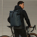 Back view of a man wearing The Roll Top in Tasmin Blue while holding onto a bike.