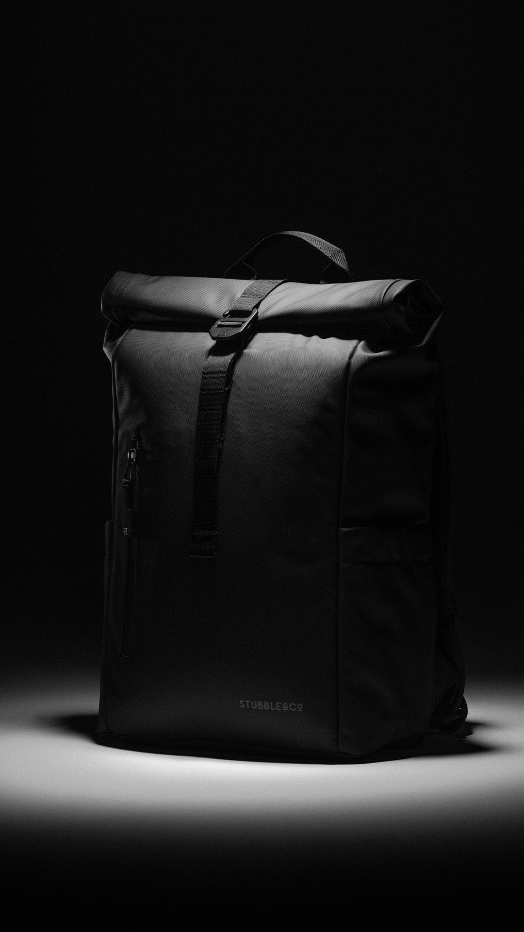 A black roll-top backpack with a front buckle strap, shown on a dark background with dramatic spotlighting.