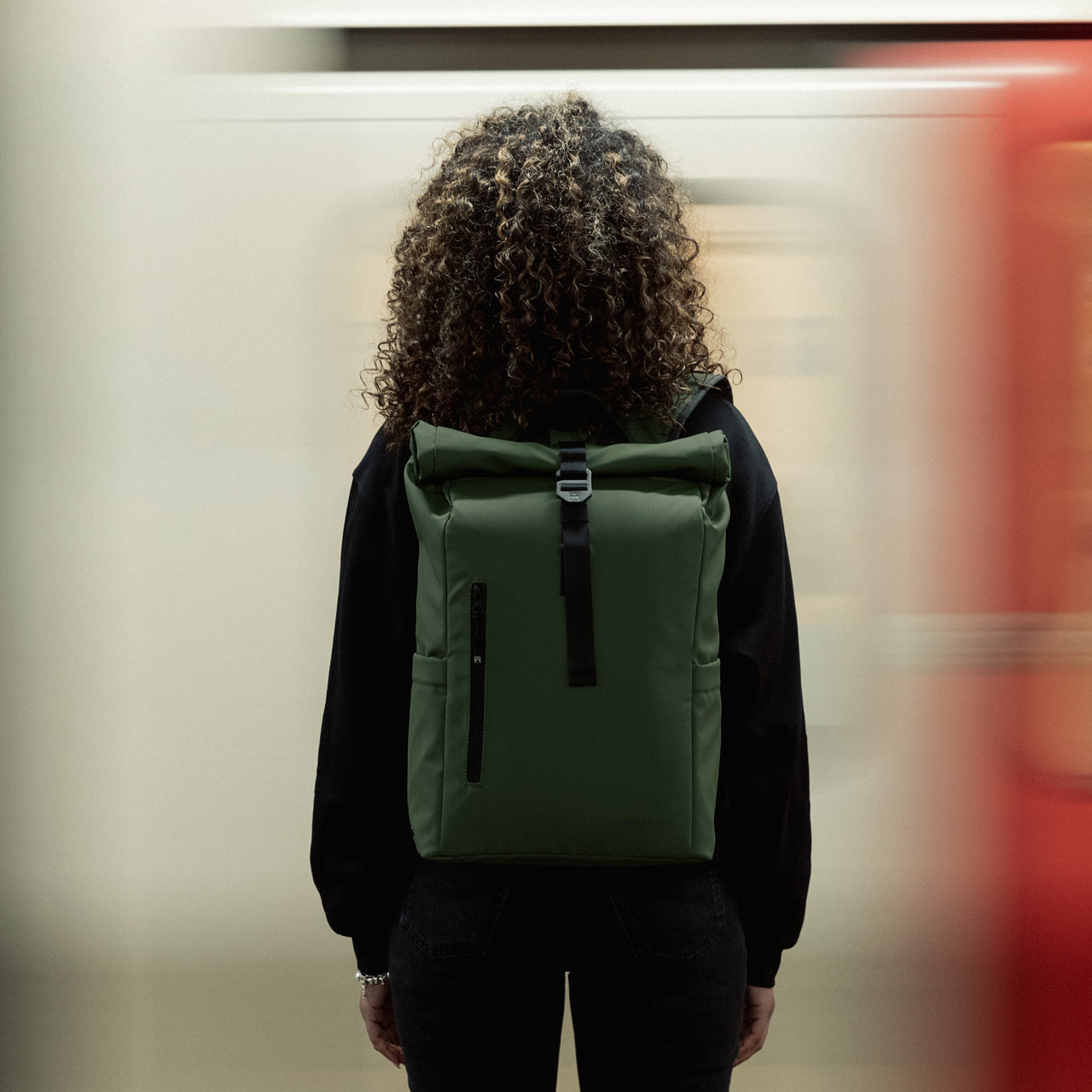 Women wearing the Roll Top 15L in Urban Green by a blurry train that is moving very fast
