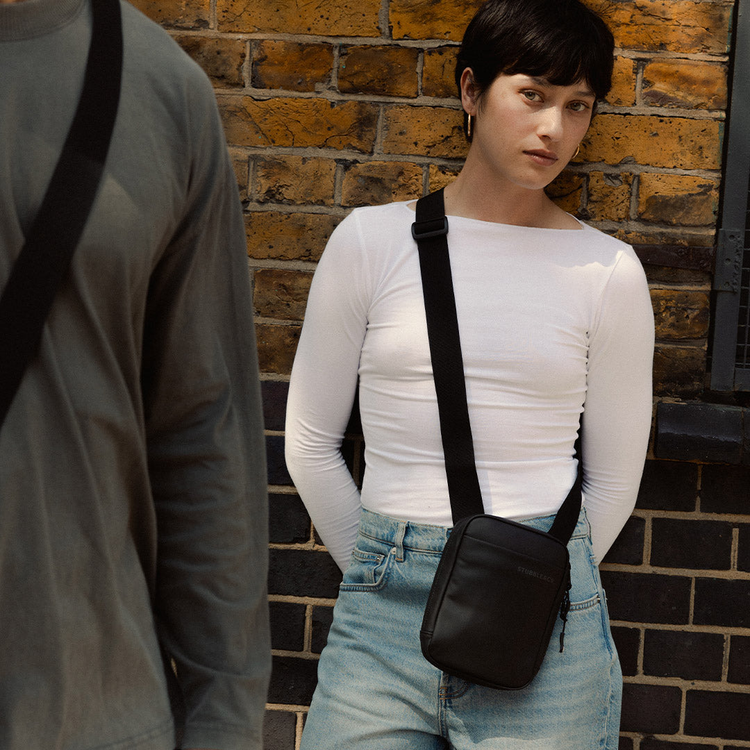 A woman standing against a wall wearing an All Black shoulder bag across her