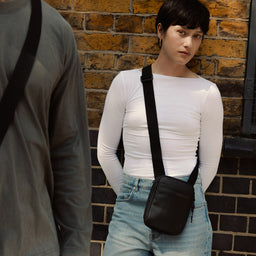 A woman standing against a wall wearing an All Black shoulder bag across her