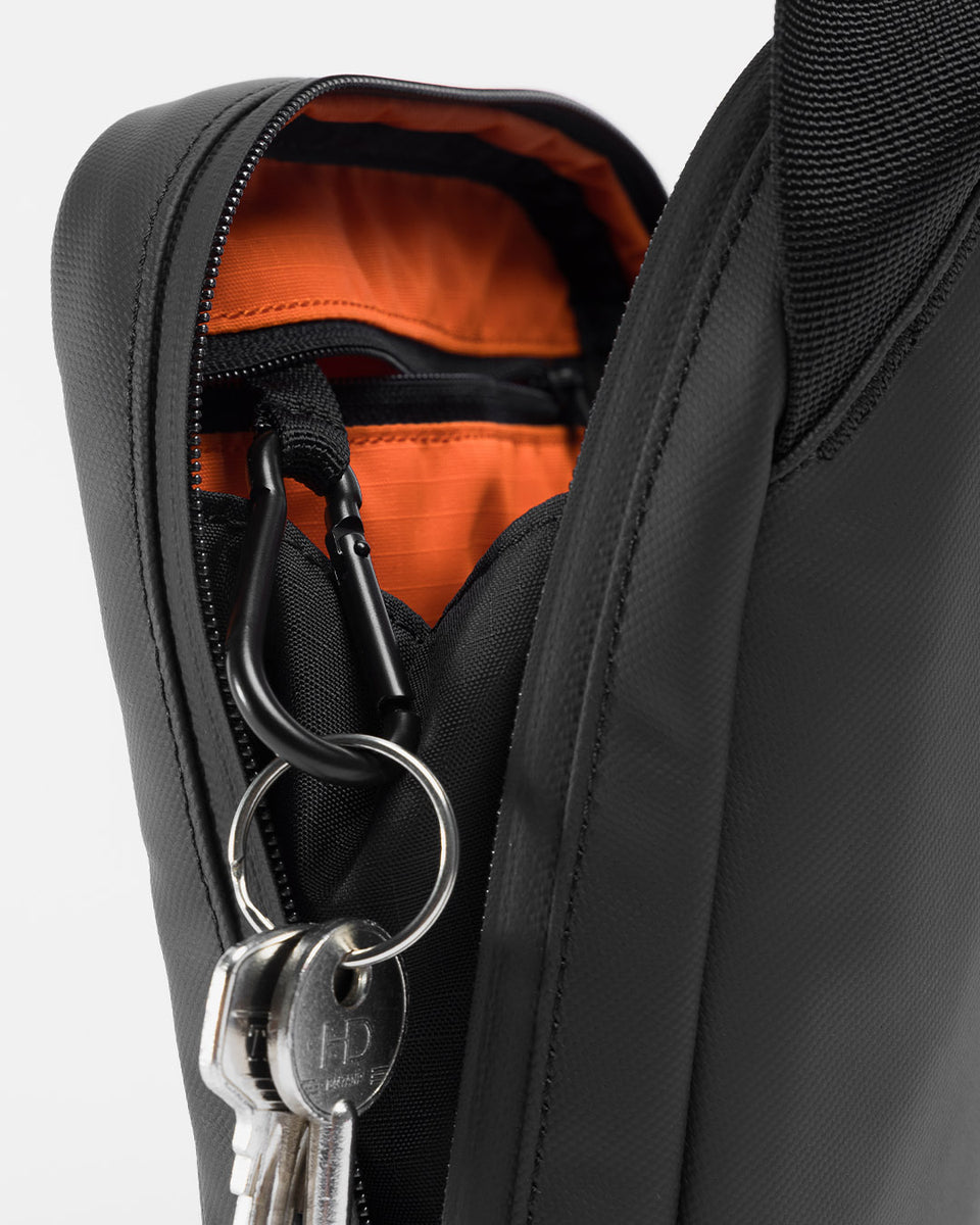 A studio shot of an all black shoulder bag showing the removable carabiner clip inside