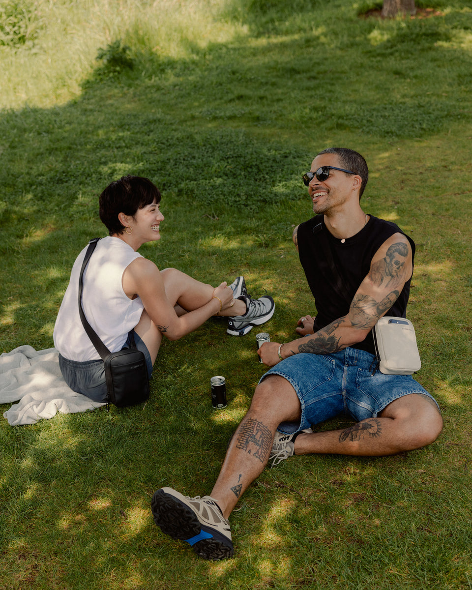 A man and woman sitting on the grass talking whilst wearing Shoulder Bags across their bodies