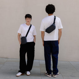 A woman wearing a Midnight Blue shoulder bag on her front standing next to a man wearing a crossbody on his back