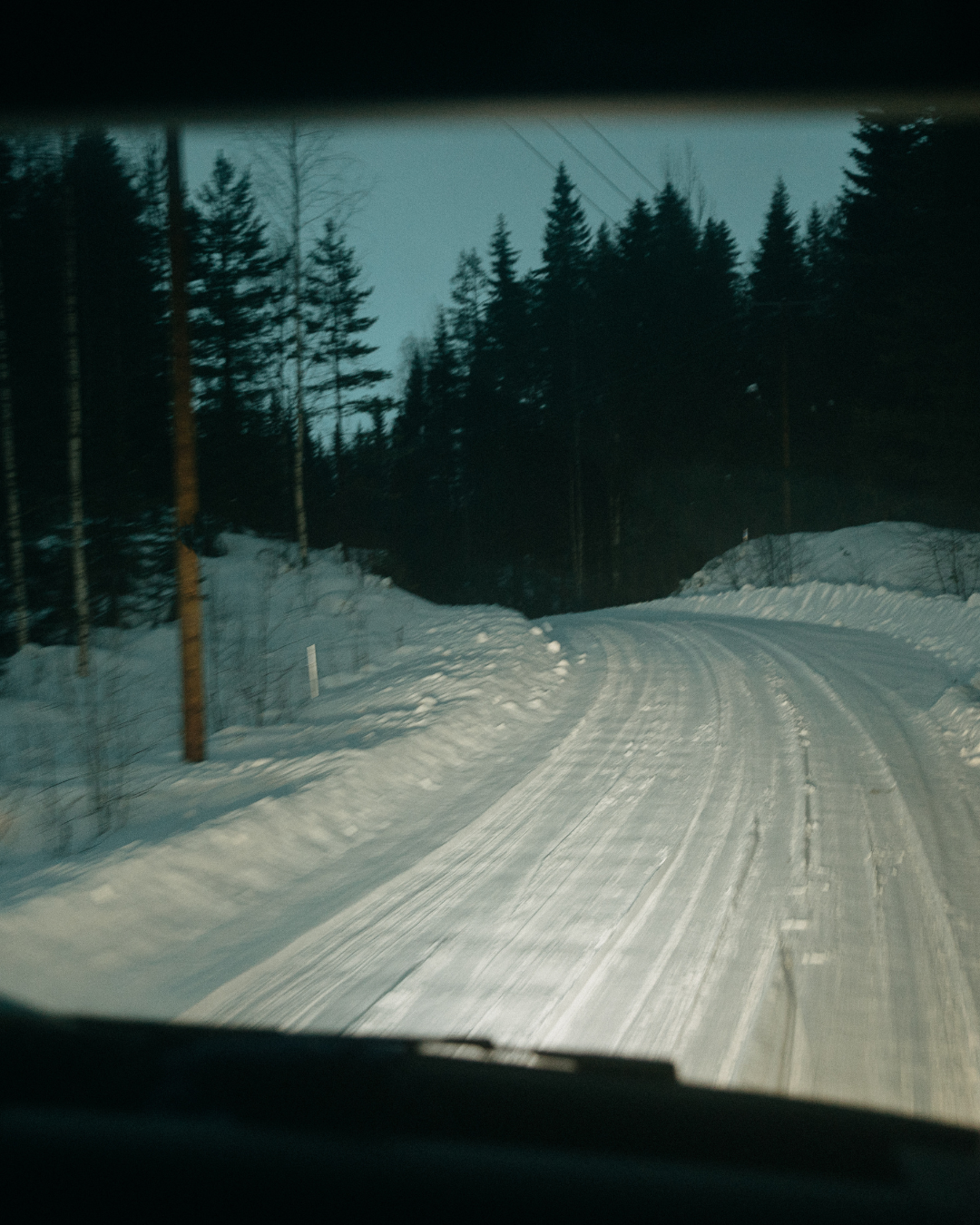 Snow-covered road seen through a car windscreen at dusk, with trees lining both sides.
