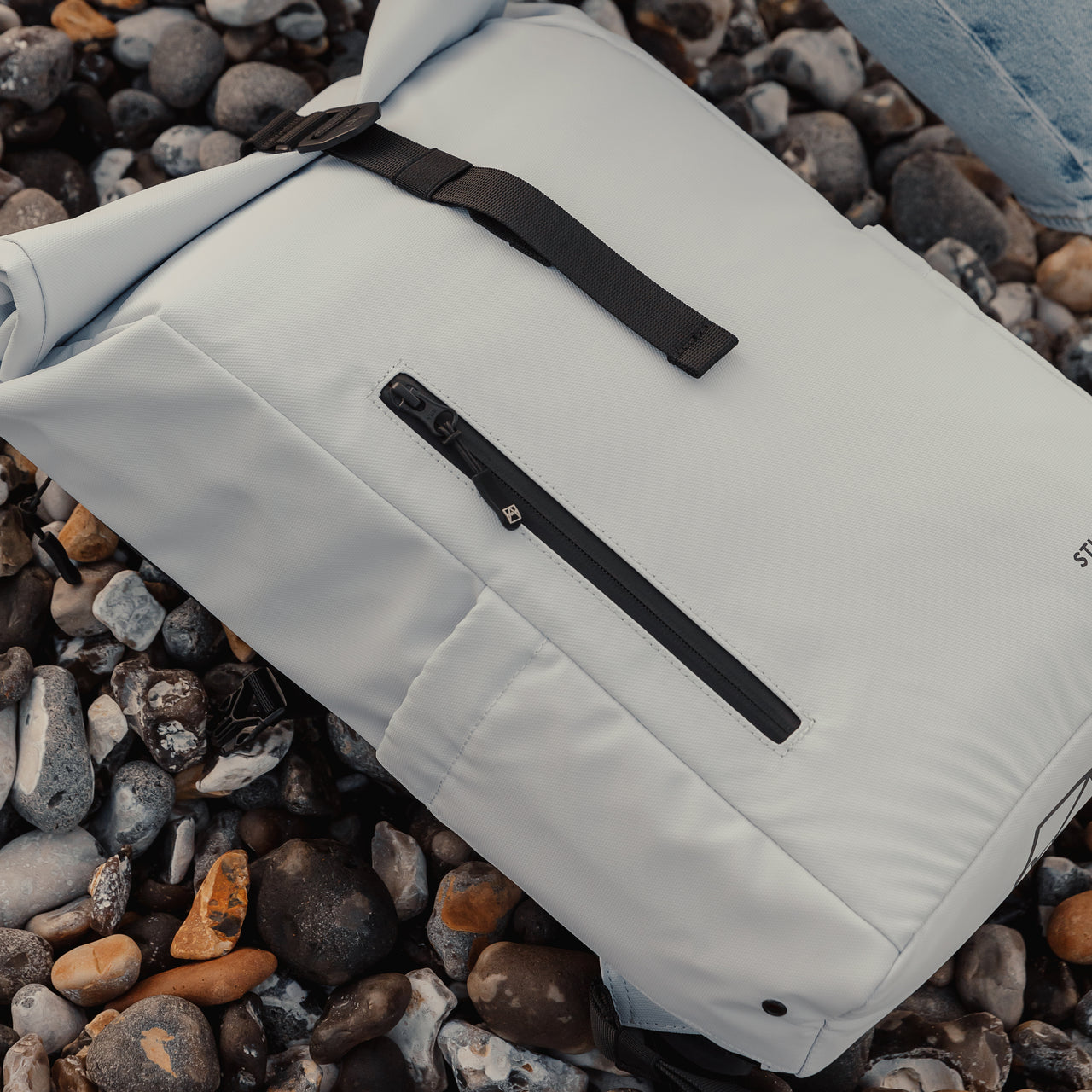 Arctic White Roll Top Mini backpack close up on a pebble beach