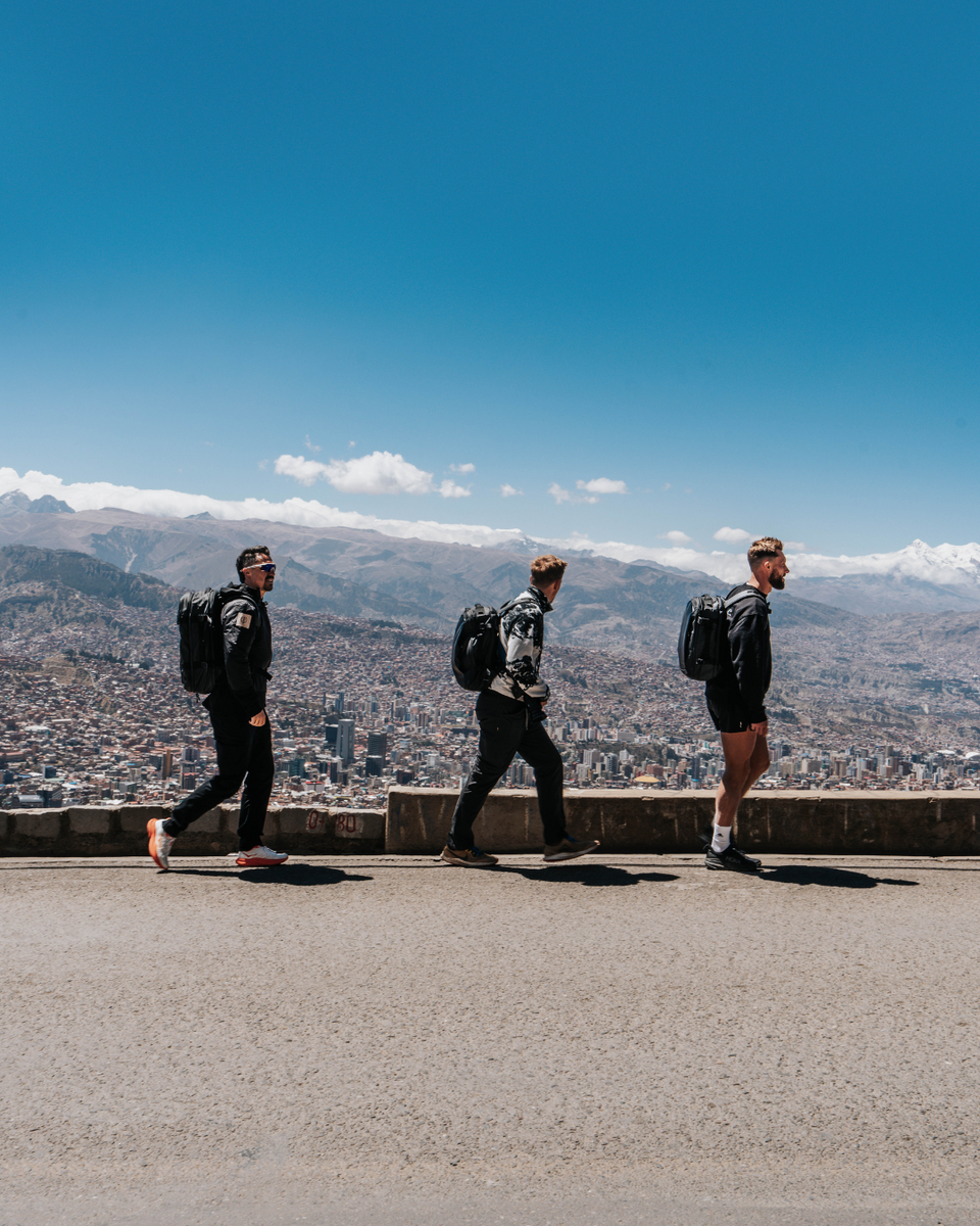 Three people with Stubble & Co backpacks walking on a road with a cityscape and mountains in the background.