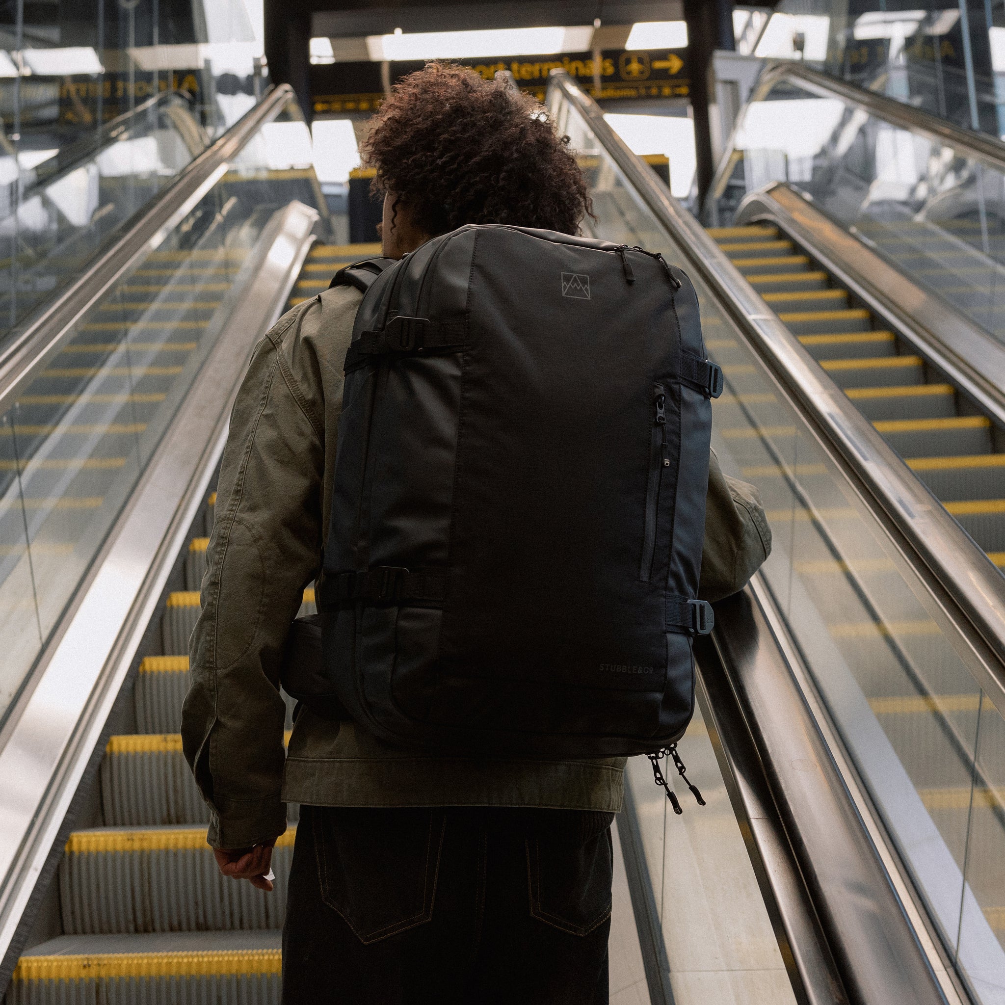 A man going up an escalator with an All Black Travel Backpack 40L on his back