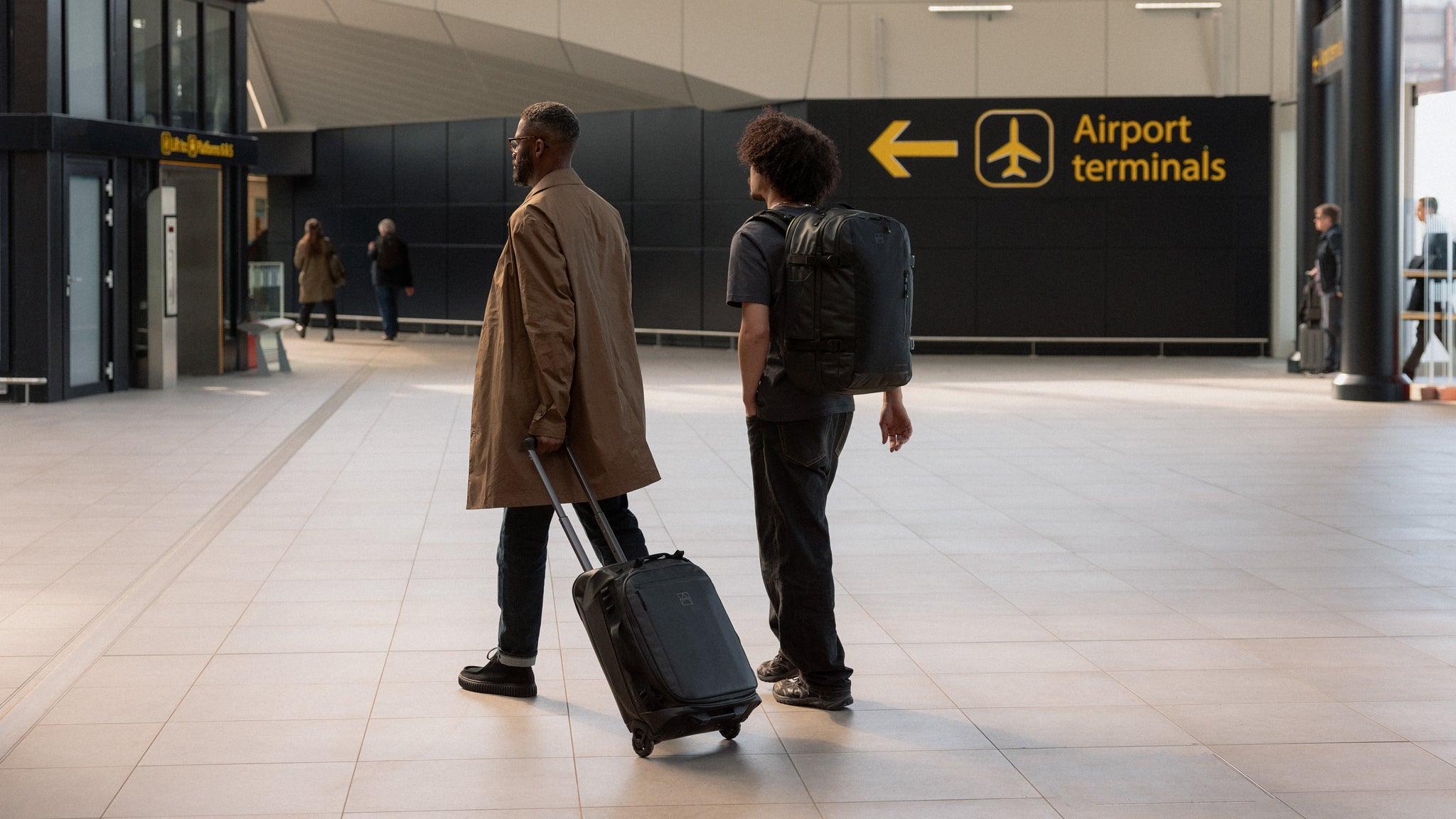 two men walking into airport departures with a wheeled kit bag 40l and travel backpack 40l