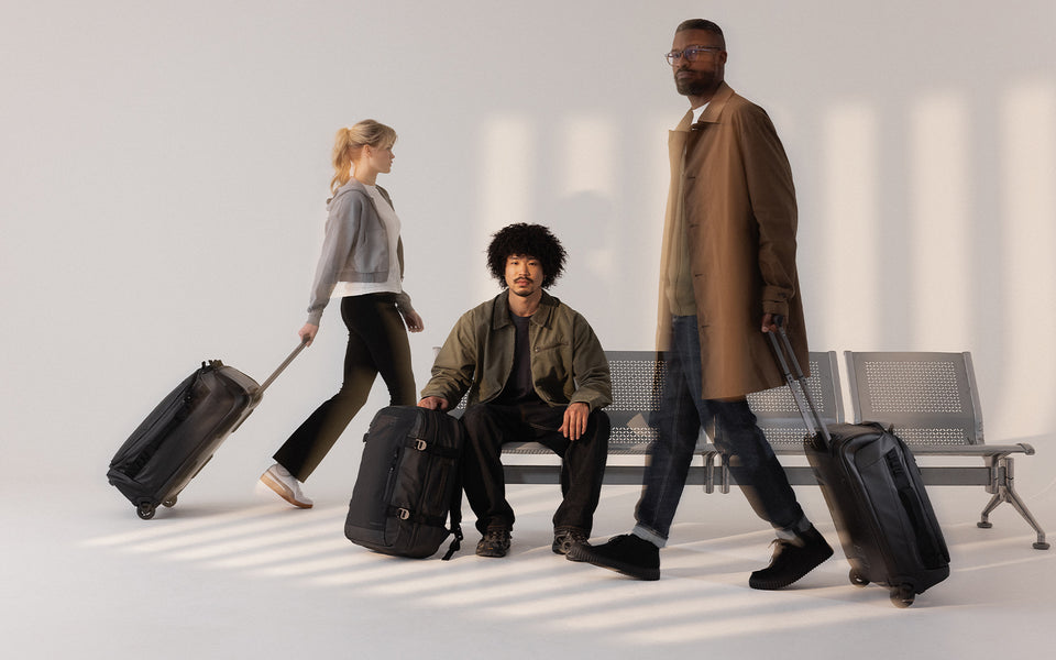 A studio with an airport chair with models walking with their luggage including the travel backpack 40l, wheeled kit bag 40l and wheeled kit bag 80l in Black