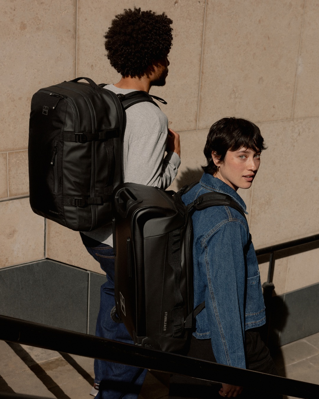 Two friends walking down steps, wearing All Black travel bags, the man with a Travel Backpack 40L and the woman with a Wheeled Duffel 40L worn as a backpack.