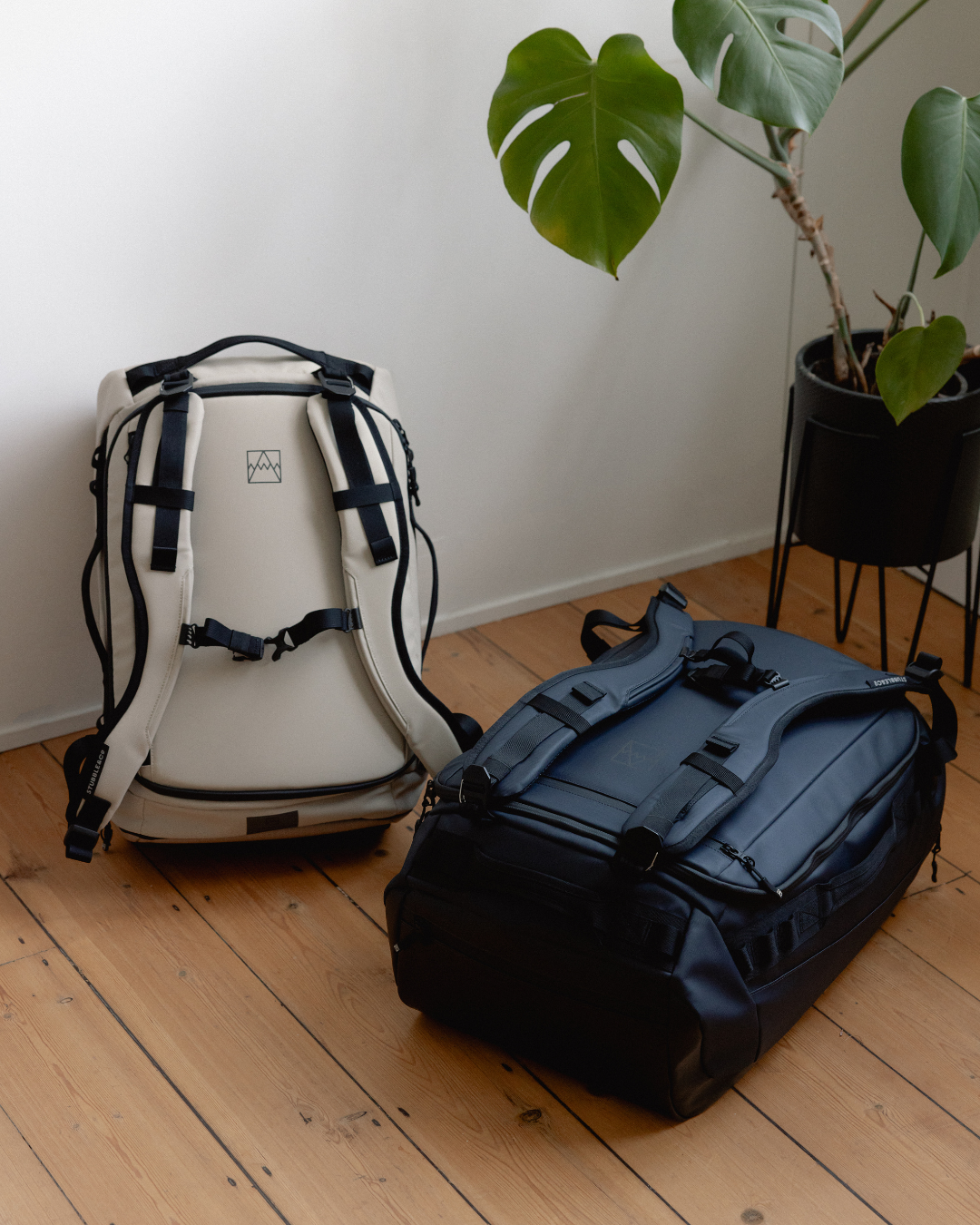 Two Kit Bags on a wooden floor, the Sand version standing upright and the Midnight Blue version lying flat, beside a potted plant. 