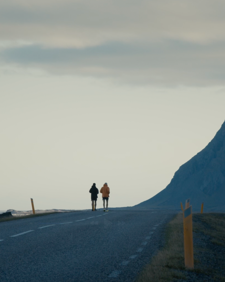 Two men run along road into the distance against a gloomy sky and mountain backdrop