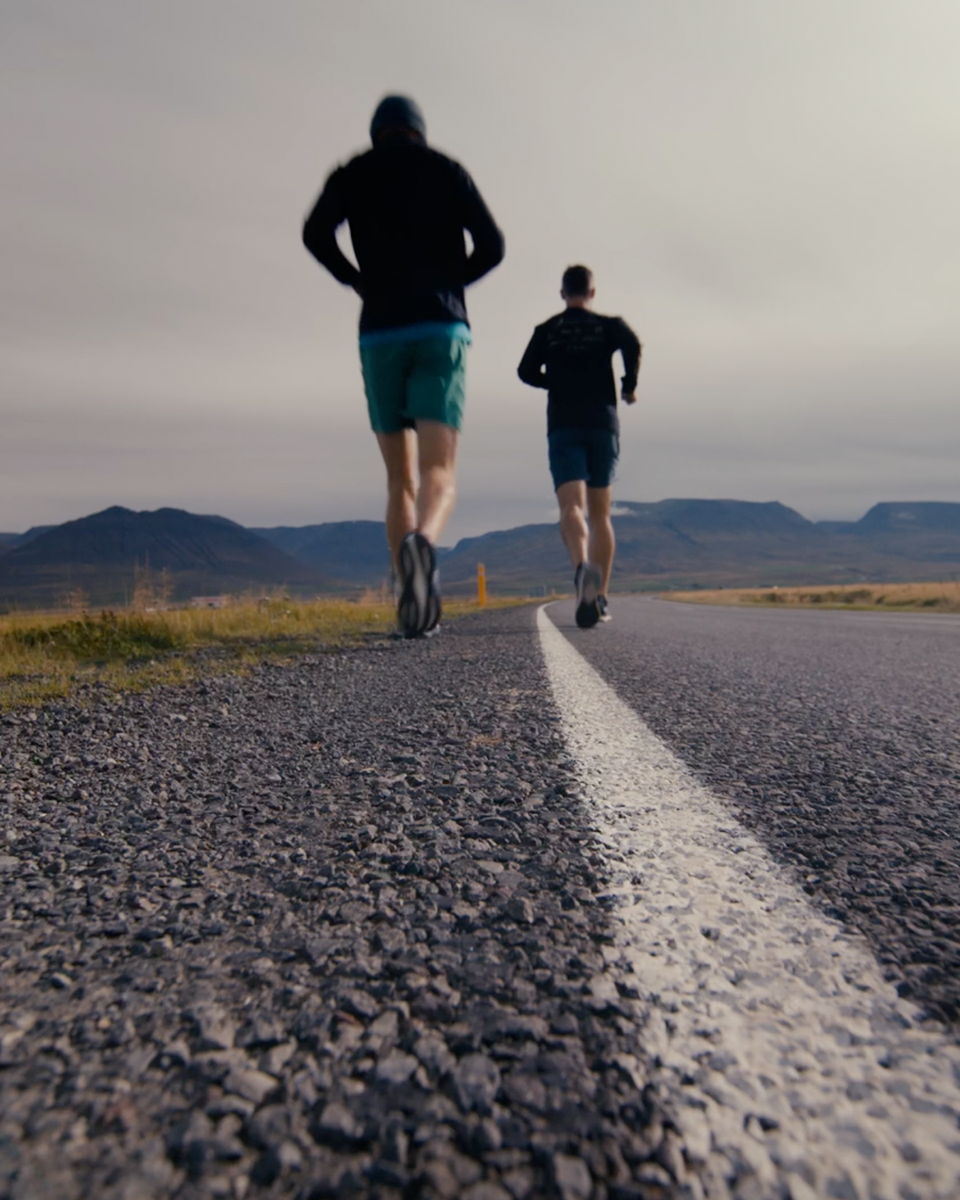 Two men run along a road away from the camera surrounded by grassy terrain and a mountain backdrop