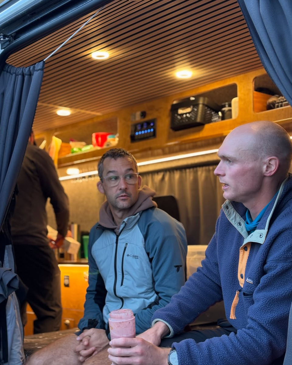 Two men drink pink smoothies whilst sat in the back of an open van chatting