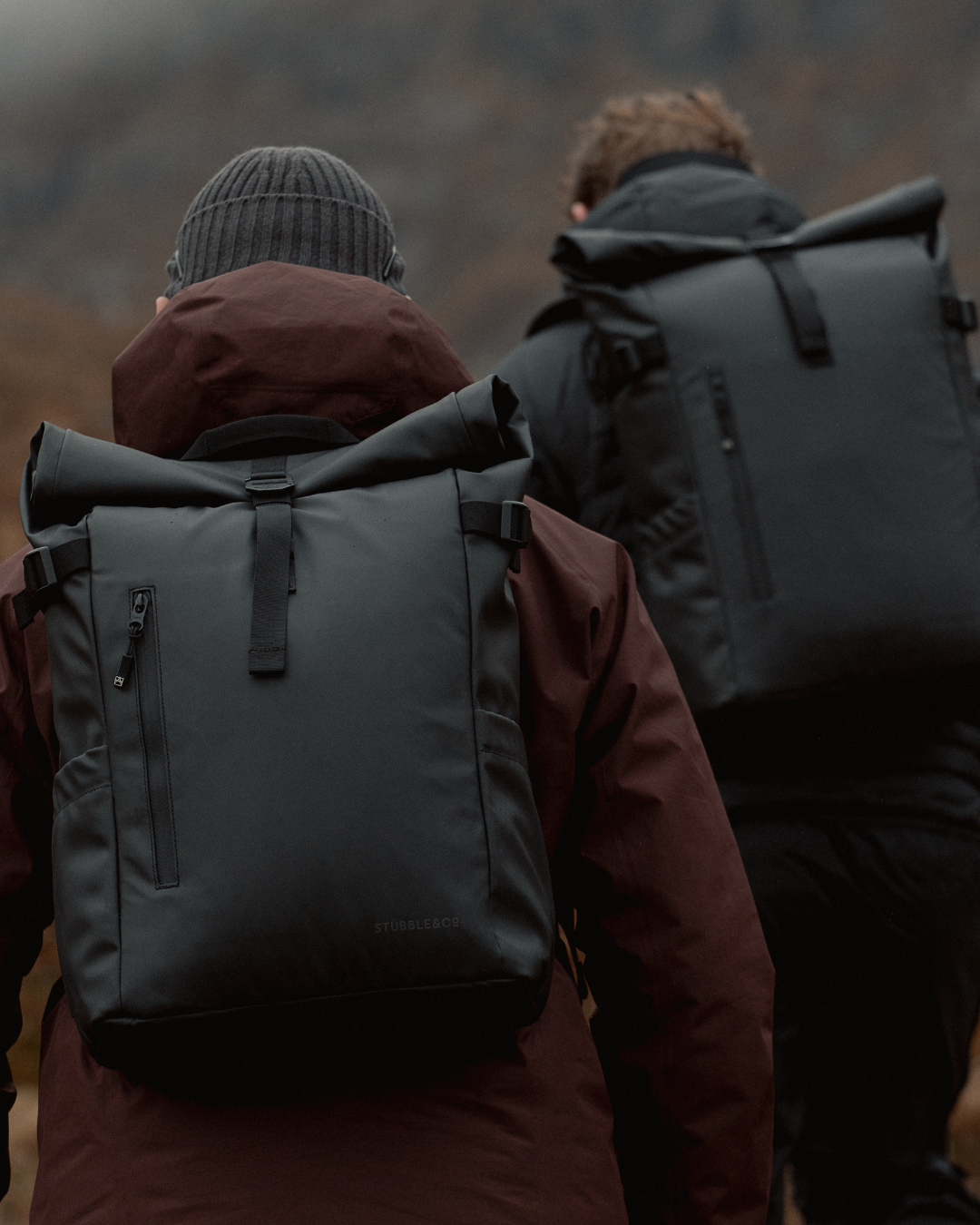 Two men walking together outside, wearing rain jackets and All Black Roll Top backpacks.
