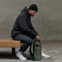 A man sitting on a bench rolling his Urban Green Roll Top backpack