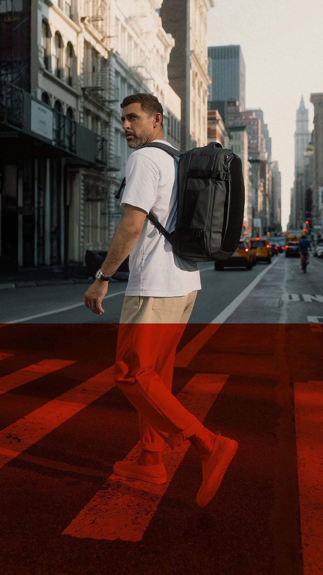 A man walking across a city crosswalk wearing a white t-shirt, beige pants, and a black backpack, with a strong red overlay across the lower half of the image.