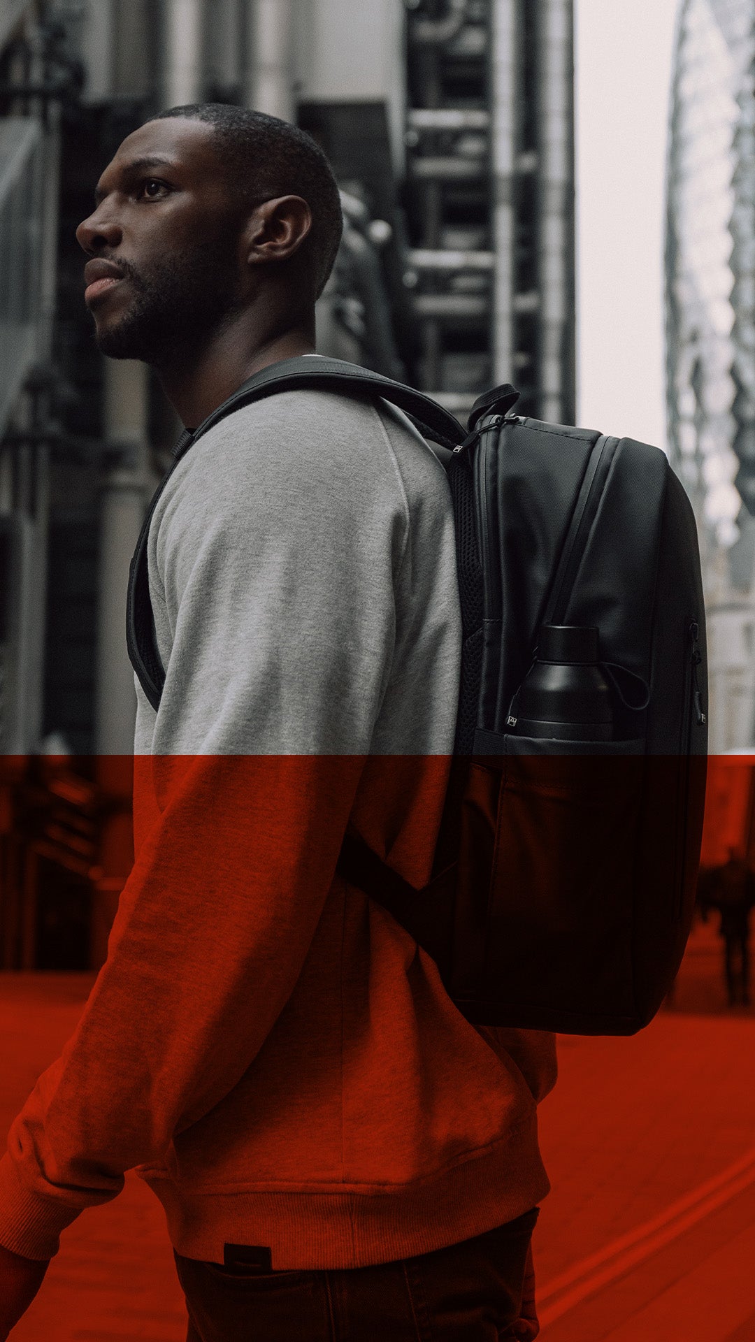 A man in a gray and red sweatshirt wearing a black backpack, captured in a city setting with a strong red overlay across the lower half of the image.