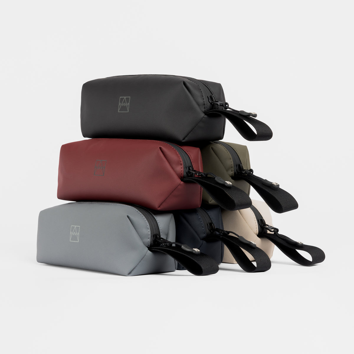 A stack of Wash Bag Minis in a range of colourways including All Black, Garnet Red and Sand
