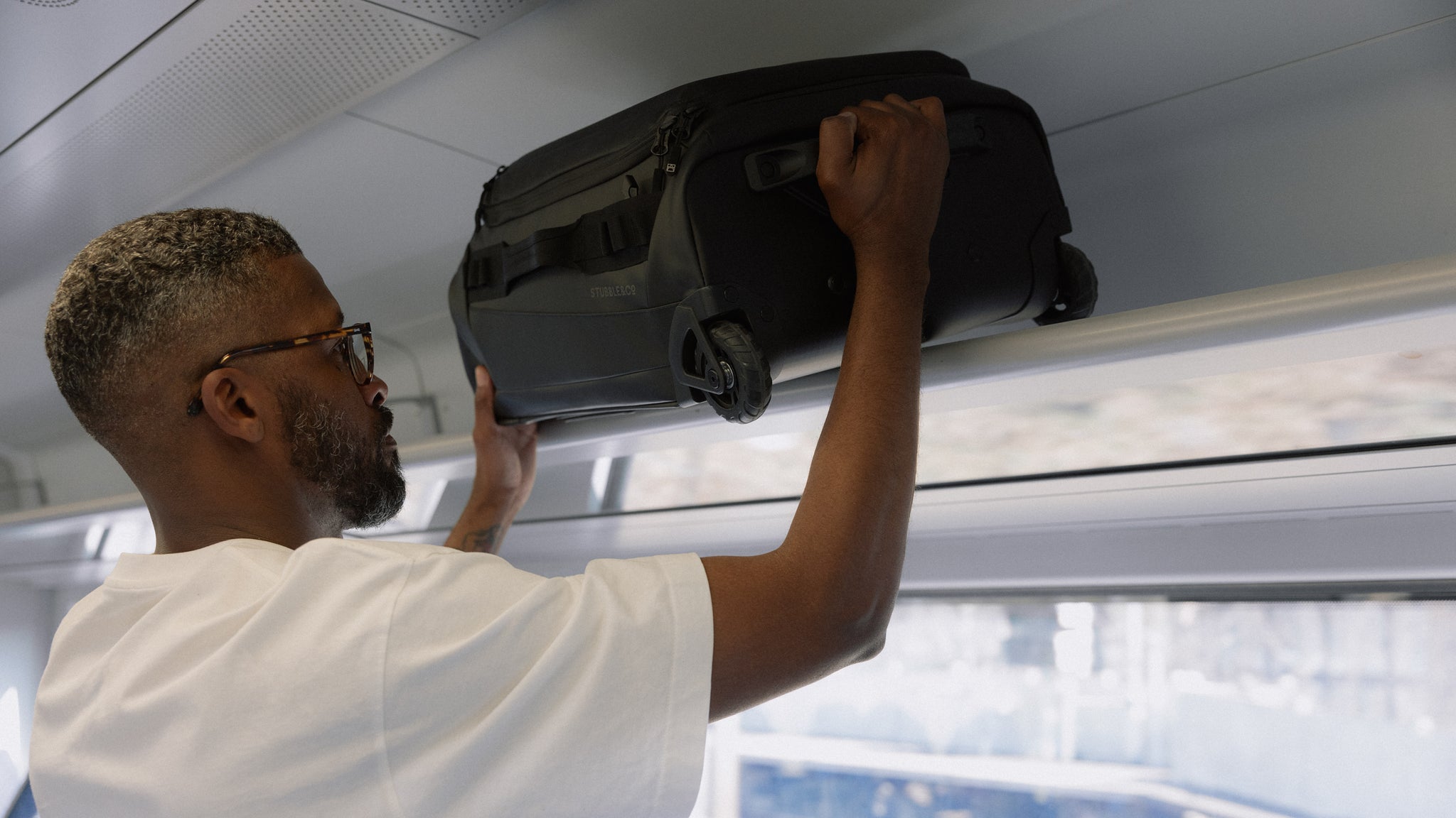 A man placing a wheeled kit bag 40l in the overhead space above a train seat