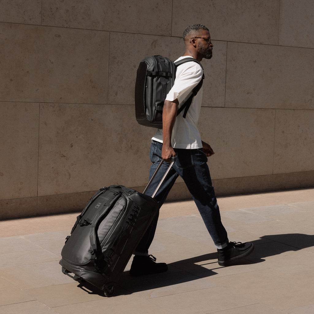 A man walking wheeling a Wheeled Duffel 80L behind him whilst wearing a Hybrid Backpack on his back. Both bags are in All Black