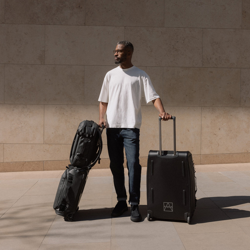 A man standing his Wheeled Duffel 80L and 40L in black with a Hybrid using the trolley sleeve