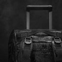 A studio shot of the All Black Wheeled Duffel 80L in All Black.  A close up of the top grab handle and telescopic handle with water on the waterproof material