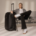 A woman sitting on an airport bench with a Wheeled Duffel 80l in All Black by her side with the telescopic handle extended
