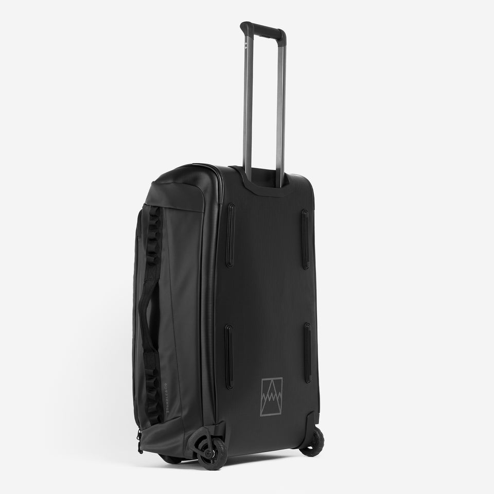 A studio shot of the All Black Wheeled Duffel 80L in All Black. it shows the back panel, reflective logo and telescopic handle