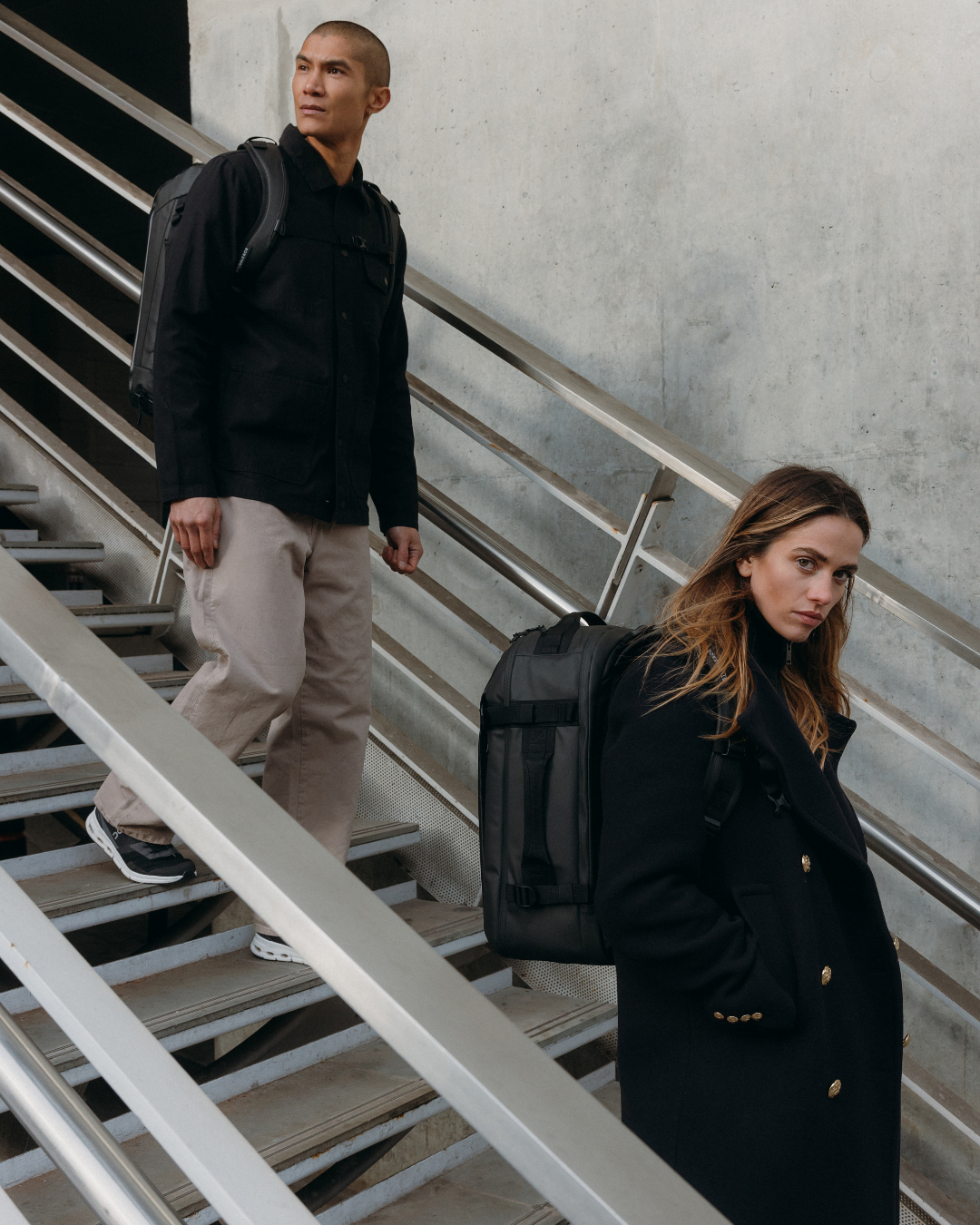 Man and woman walking down stairs wearing casual outfits and carrying hybrid backpacks.