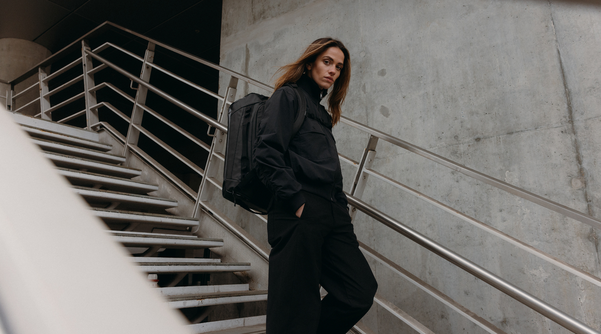 Woman in a black jacket walking down stairs wearing the Kit Bag in All Black on her back. 