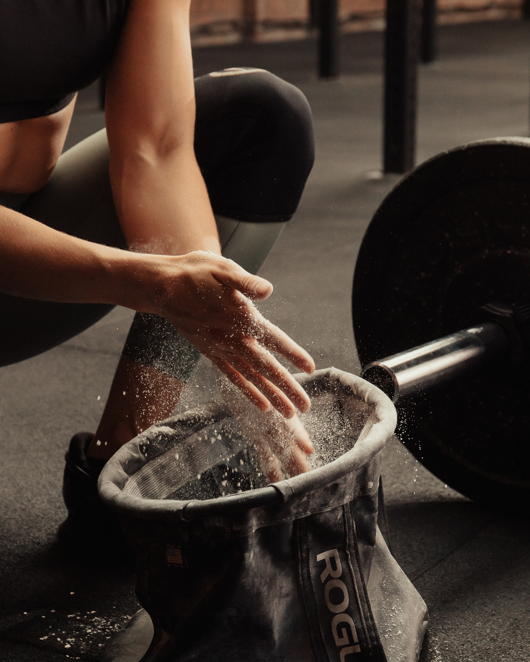 A woman crouching at the gym, dipping her hand into a chalk bag on the floor beside a barbell.