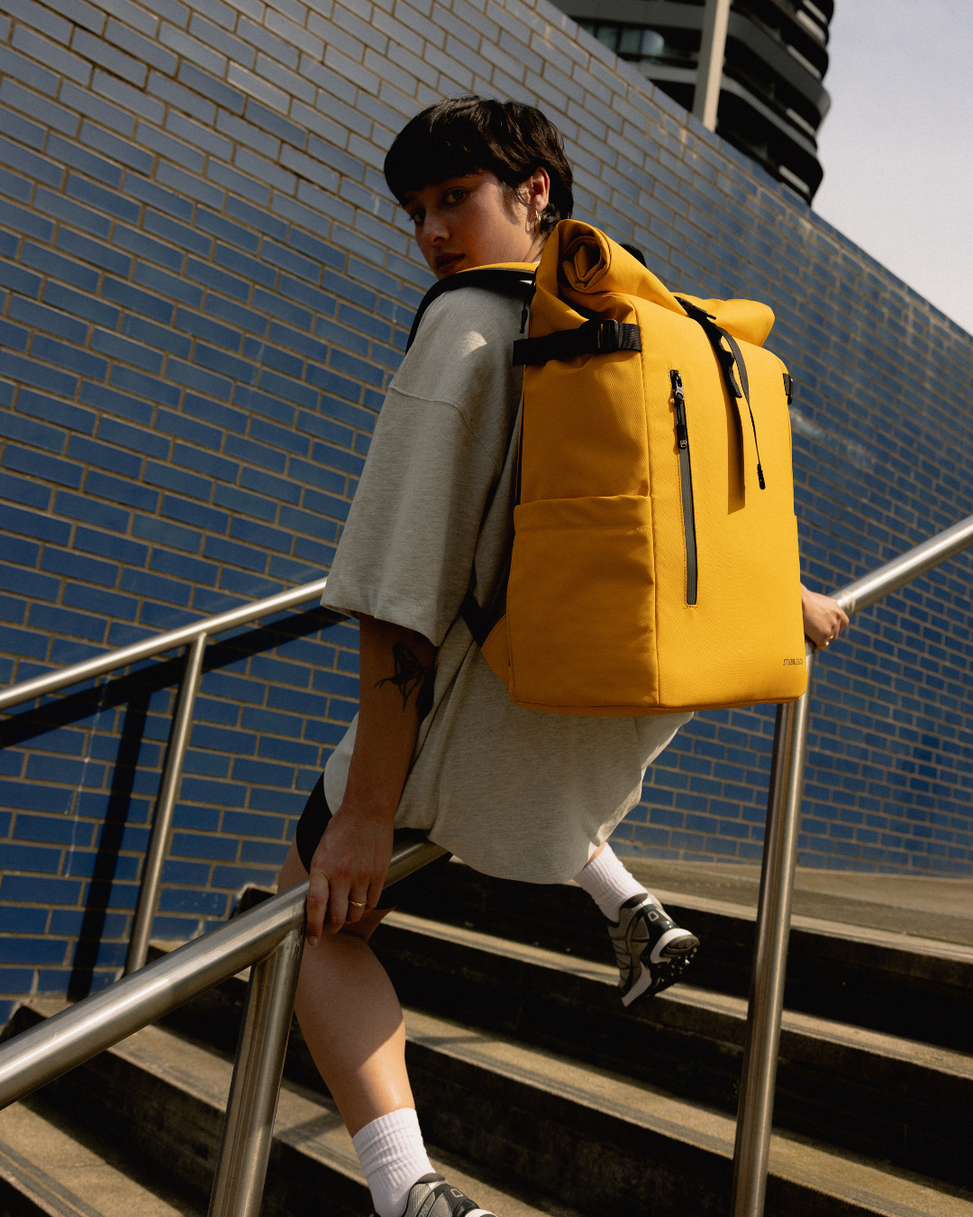 A woman sitting on a railing, wearing a Roll Top Backpack in Yellow.