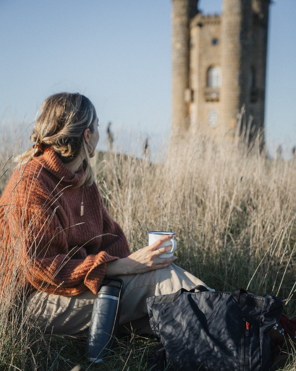 a woman sitting with a mug in front of a castle with a black packable tote - the travel project blog