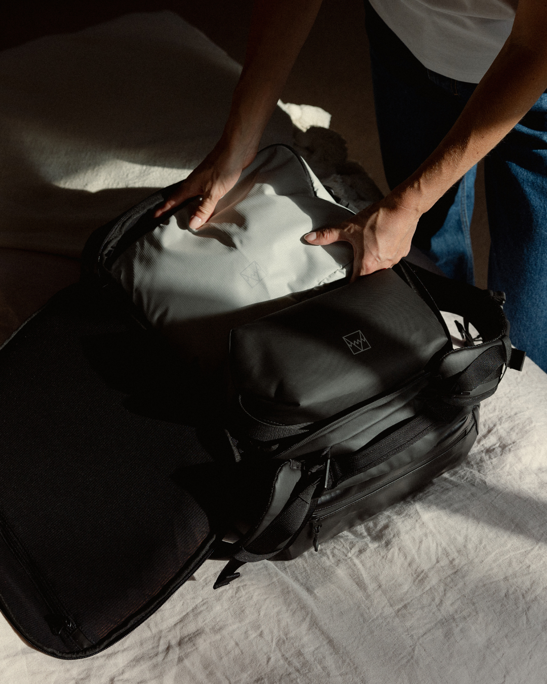 A person's hands unpacking an All Black Kit Bag on a bed, revealing an Off-White Packing Cube and an All Black Wash Bag.