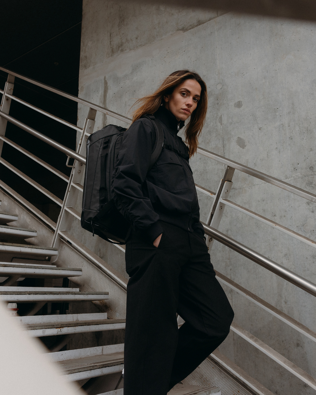 A woman wearing a black outfit walking down a staircase, wearing an All Black Kit Bag on her shoulders.