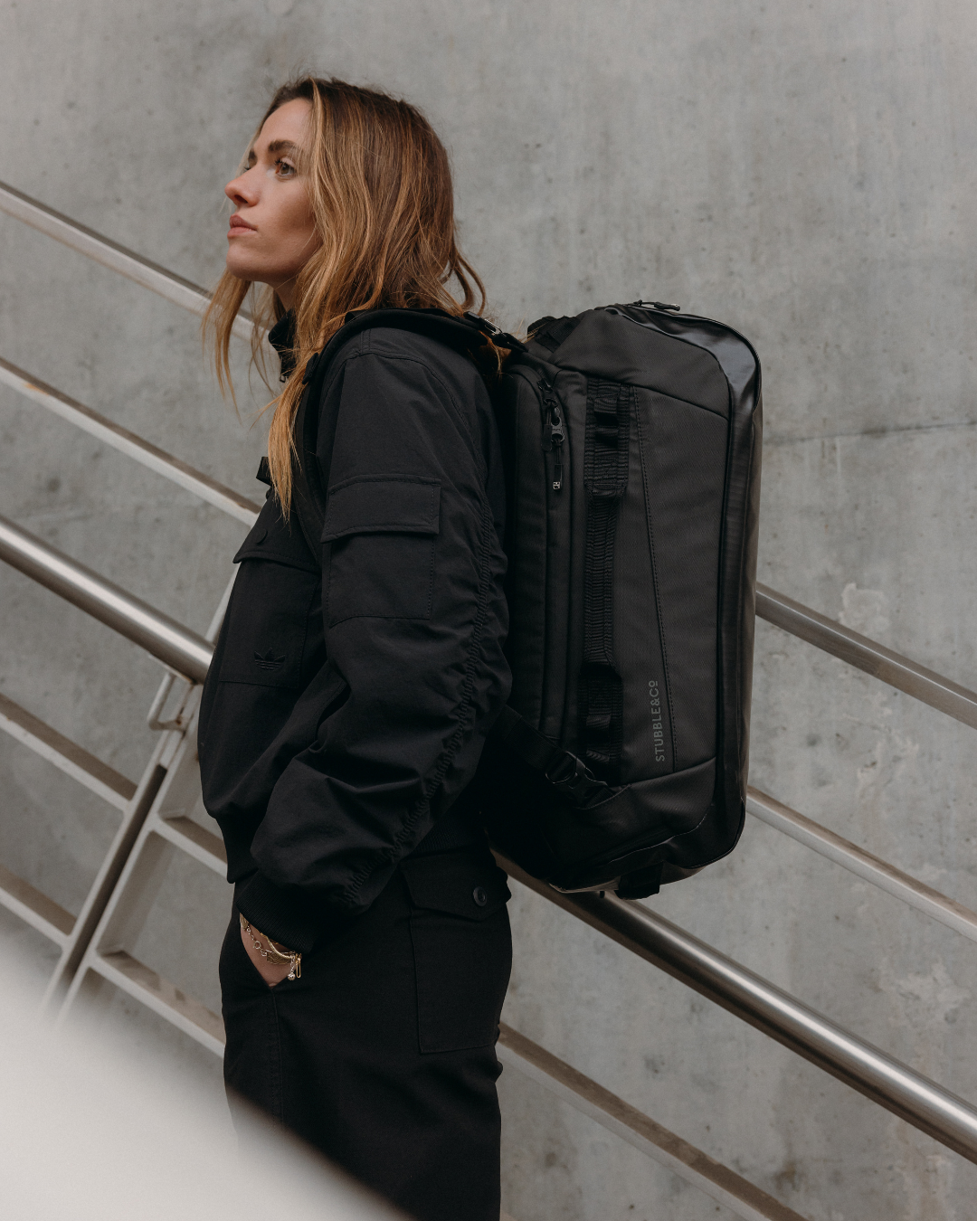 Woman in a black jacket walking upstairs wearing the Kit Bag in All Black. 