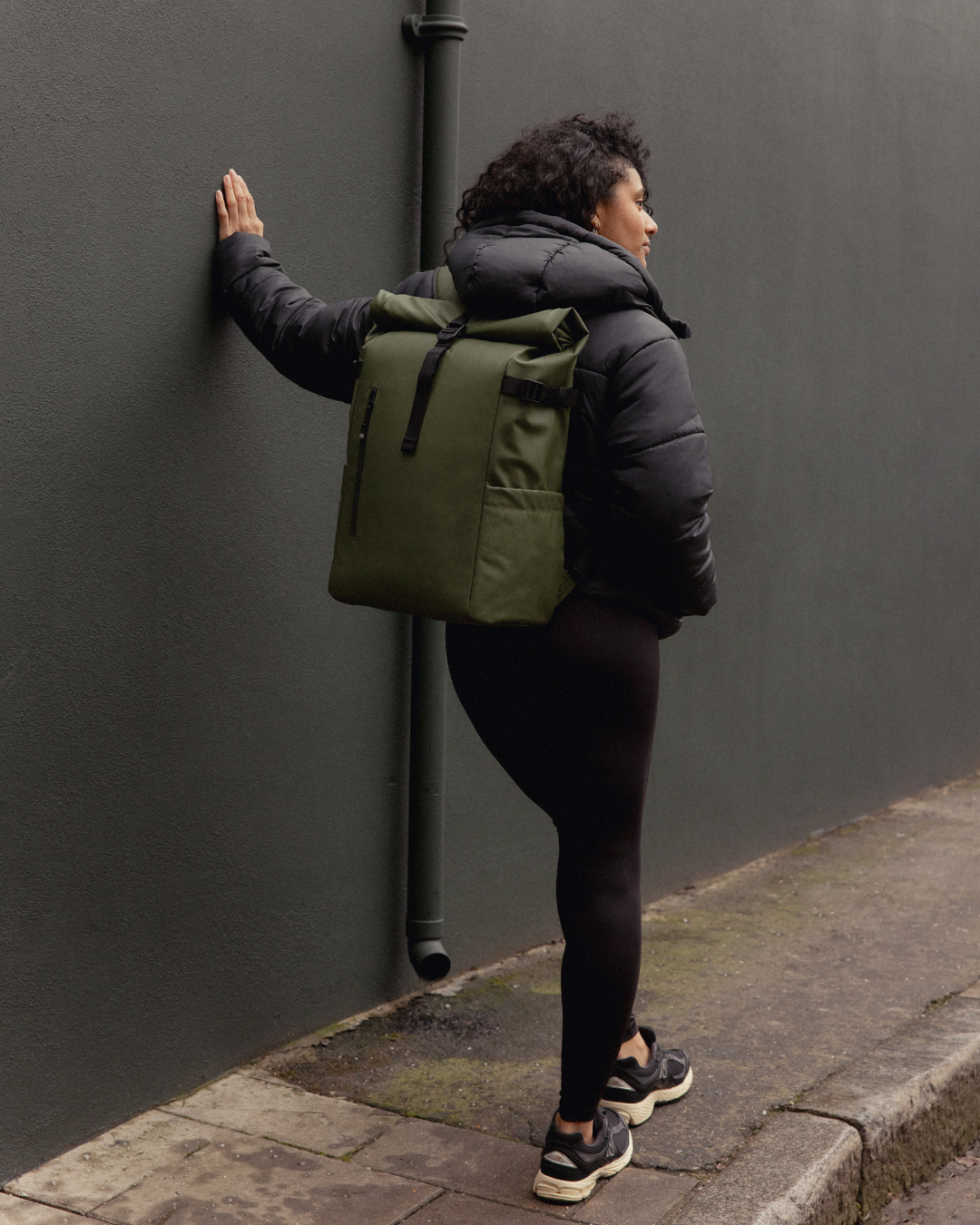 A woman wearing a black puffer jacket and a Roll Top Backpack in Urban Green, on a London street.
