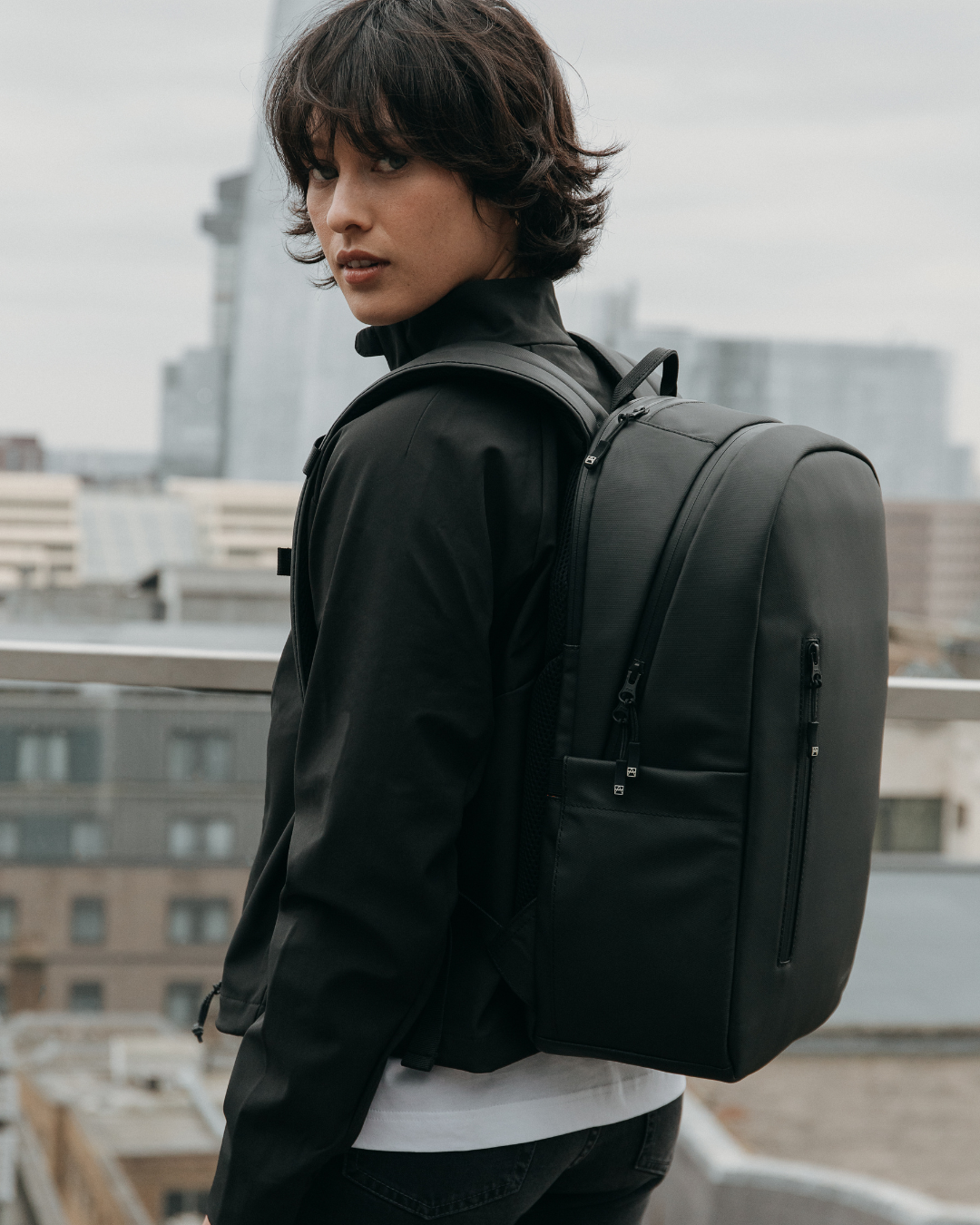 Woman in a black jacket wearing the Everyday Backpack in All Black, with a city skyline behind her.