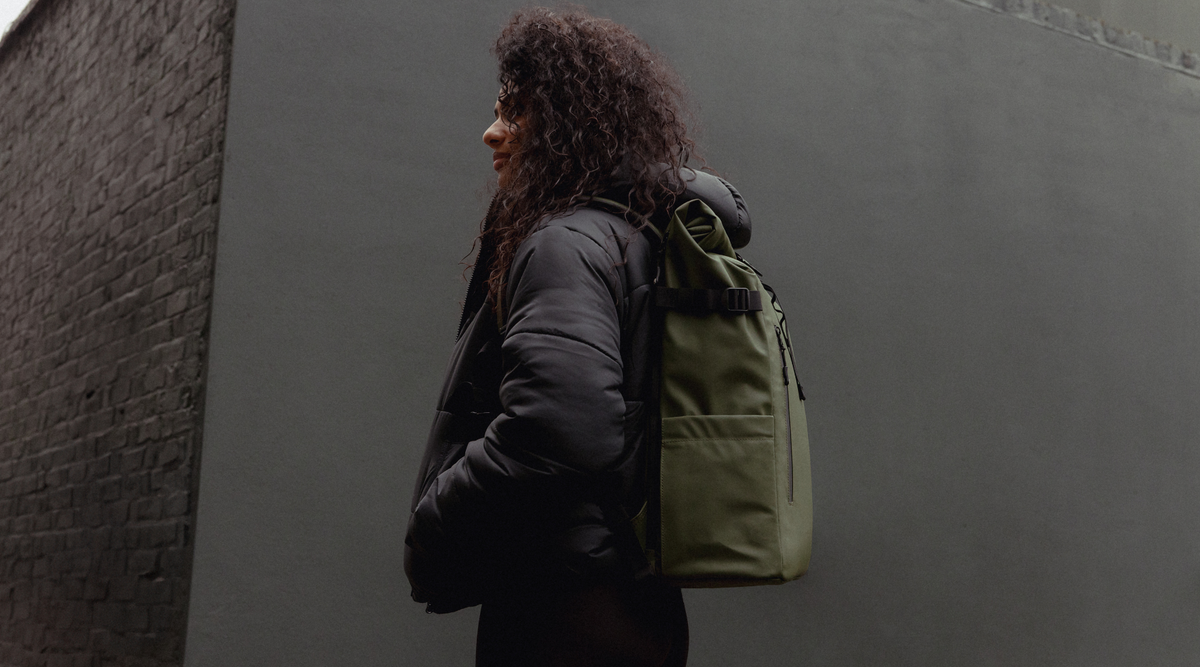 A woman wearing a black puffer jacket, carrying a Roll Top Backpack in Urban Green.