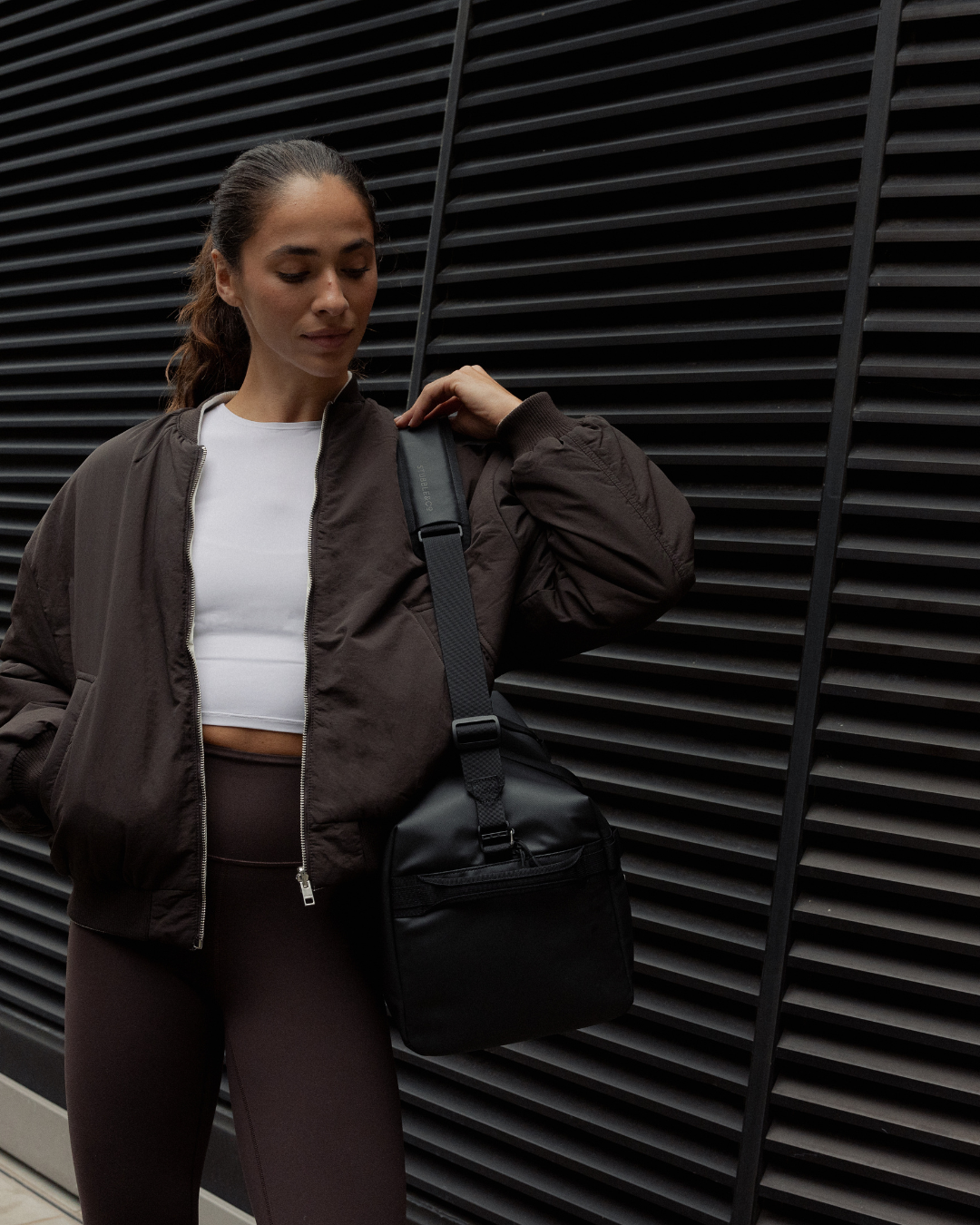 A woman wearing an All Black Gym Duffel over her shoulder, dressed in a brown bomber jacket.