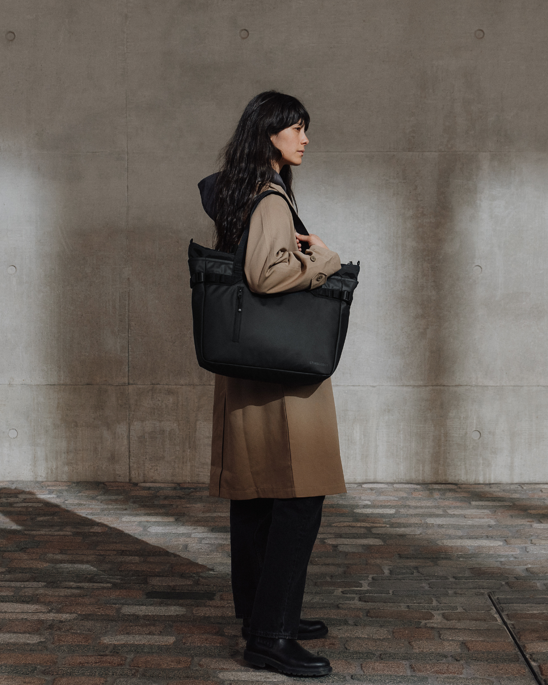 Woman in a beige trench coat carrying the Laptop Tote 20L in All Black against a concrete wall.